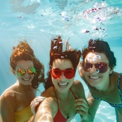 Rambler:Cheerful women friends swimming underwater in pool taking selfie. Underwater selfie of happy females in pool.